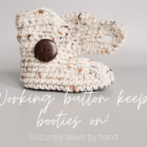 May include: Cream-colored crocheted baby booties with a brown wooden button. The text "Working button keeps booties on! Securely sewn by hand" is below the booties.