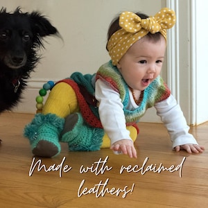 May include: A baby wearing a yellow polka dot headband, a colorful knitted vest, and turquoise booties. The text "Made with reclaimed leathers!" is written in a script font. A black dog is in the background.