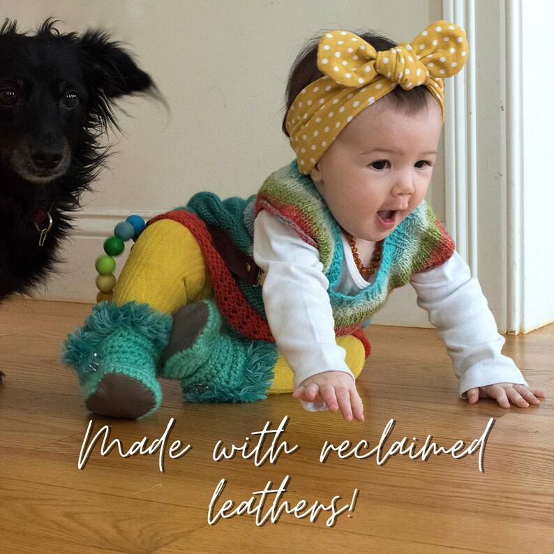 May include: A baby wearing a yellow polka dot headband, a colorful knitted vest, and turquoise booties. The text "Made with reclaimed leathers!" is written in a script font. A black dog is in the background.