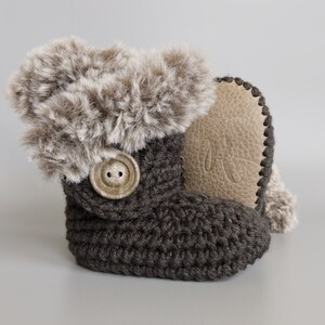 May include: A single dark gray crochet baby bootie with a faux fur trim and a large button closure. The bootie has a light brown leather sole with an embossed letter. The bootie is designed for infants.