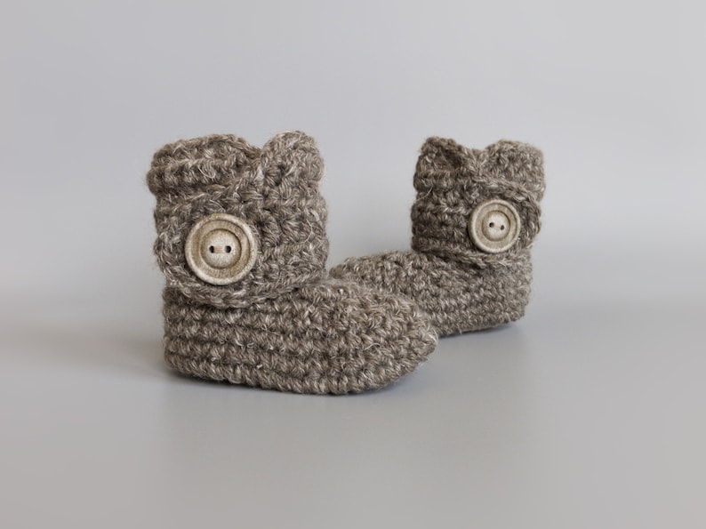 May include: A pair of brown crocheted baby booties with a button on each. The booties are made with a soft, textured yarn.