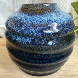 以下が含まれることがあります： 丸みを帯びたボディと広い開口部を持つ、手作りのセラミック花瓶。 青、黒、茶色の色合いで、テクスチャ加工されたレイヤードデザインが特徴です。 光沢のある仕上がりは色の深みを増し、装飾的な作品となっています。