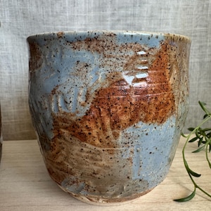 May include: Handmade ceramic planter with a wide, rounded shape. The exterior features a blend of blue, brown, and tan hues with a speckled texture. The planter has a rustic, organic aesthetic, suitable for indoor or outdoor use.