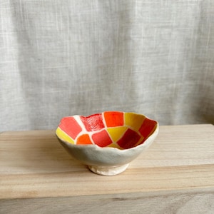 Op de afbeelding: Een kleine keramische kom met een geometrisch patroon in tinten rood, oranje en geel. De kom heeft een licht onregelmatige vorm en een klein voetje.