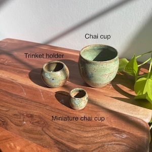 Puede incluir: Tres tazas de cerámica hechas a mano de diferentes tamaños, etiquetadas como "Chai cup", "Trinket holder" y "Miniature chai cup". Las tazas tienen un esmalte verde claro moteado con motas marrones. Están sobre una superficie de madera.