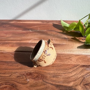 Könnte beinhalten: Eine kleine, handgefertigte Keramikschale mit beigefarbenem Äußeren und hellblauem Rand. Die Schale hat braune Blumenmuster und steht auf drei kleinen Füßen. Die Schale wird auf einer Holzoberfläche präsentiert.