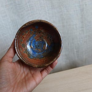 May include: A small, handmade ceramic bowl with a unique glaze. The interior features a vibrant blue center surrounded by speckled orange and brown hues. The bowl's rim is a darker brown, and it appears to be approximately 4 inches in diameter.