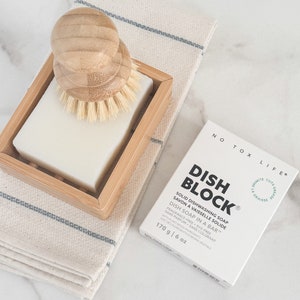 May include: A white bar of dish soap in a bamboo soap dish with a wooden brush. The soap bar packaging is white with green text that reads "Dish Block" and "Solid Dishwashing Soap". The packaging also says "No Tox Life" and "170 g | 6 oz".