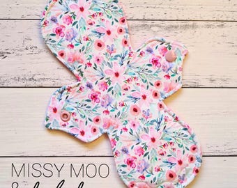 May include: A white reusable cloth pad with a pink floral print. The pad has two pink snaps on the sides. The text "Missy Moo & baby boo" is printed on the bottom of the pad.
