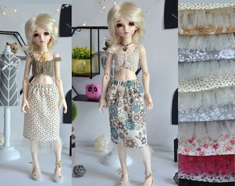 May include: Two dolls wearing different skirts and tops. The doll on the left is wearing a white skirt with brown polka dots and a brown crochet top. The doll on the right is wearing a floral skirt with a white crochet top. Both dolls are wearing a white tulle underskirt.