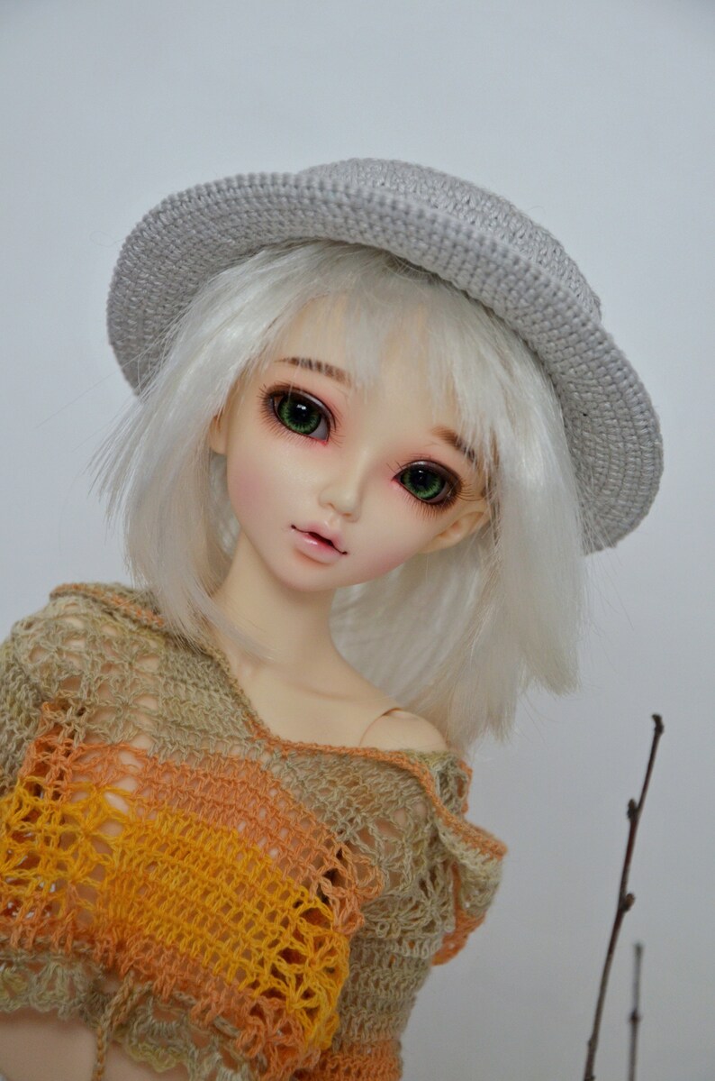 Peut inclure: Une poup&eacute;e aux cheveux blancs et aux yeux verts portant un chapeau crochet&eacute; gris et un haut crochet&eacute; multicolore.