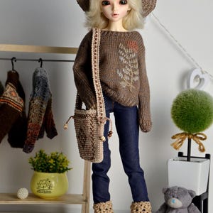 May include: A doll wearing a brown knit hat, sweater, and boots, paired with dark blue jeans and a matching purse. The sweater features an embroidered leaf design. The doll is posed in front of a decorative shelf with a small plant and a teddy bear.
