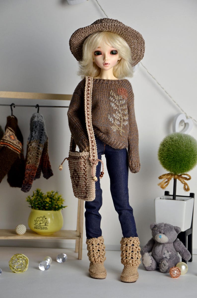 May include: A doll wearing a brown knit hat, sweater, and boots, paired with dark blue jeans and a matching purse. The sweater features an embroidered leaf design. The doll is posed in front of a decorative shelf with a small plant and a teddy bear.