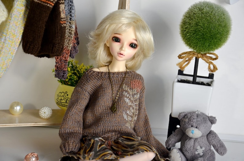 May include: A doll with blonde hair wearing a brown sweater and patterned skirt. The doll has a necklace and is posed in front of a small green topiary and a gray teddy bear. Decorative items are on a shelf behind the doll.