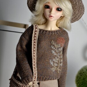 May include: A doll wearing a brown knit sweater with floral embroidery, a matching hat, and a shoulder bag. The doll has blonde hair and is wearing tan pants. The sweater features a detailed floral design.