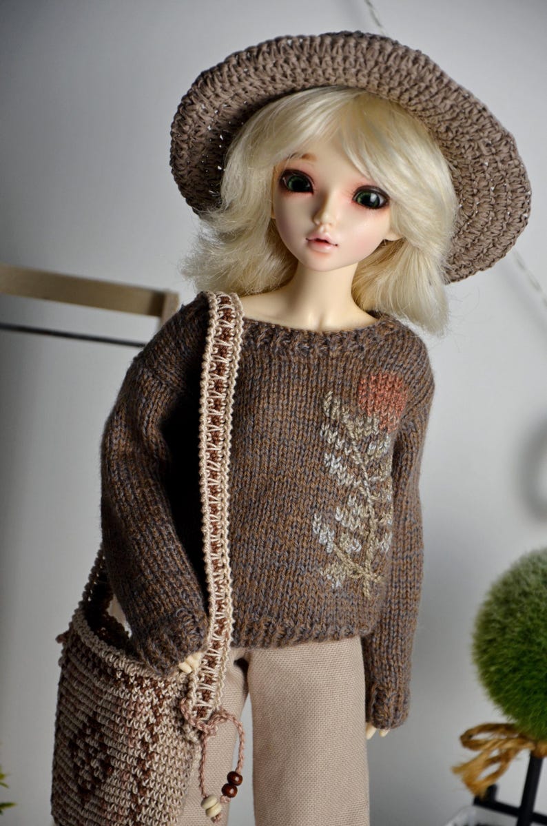 May include: A doll wearing a brown knit sweater with floral embroidery, a matching hat, and a shoulder bag. The doll has blonde hair and is wearing tan pants. The sweater features a detailed floral design.