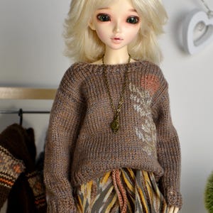 May include: A doll with blonde hair wearing a brown knit sweater with a decorative design and a patterned skirt. The doll has a long bronze necklace. The doll is standing in front of a white wall.