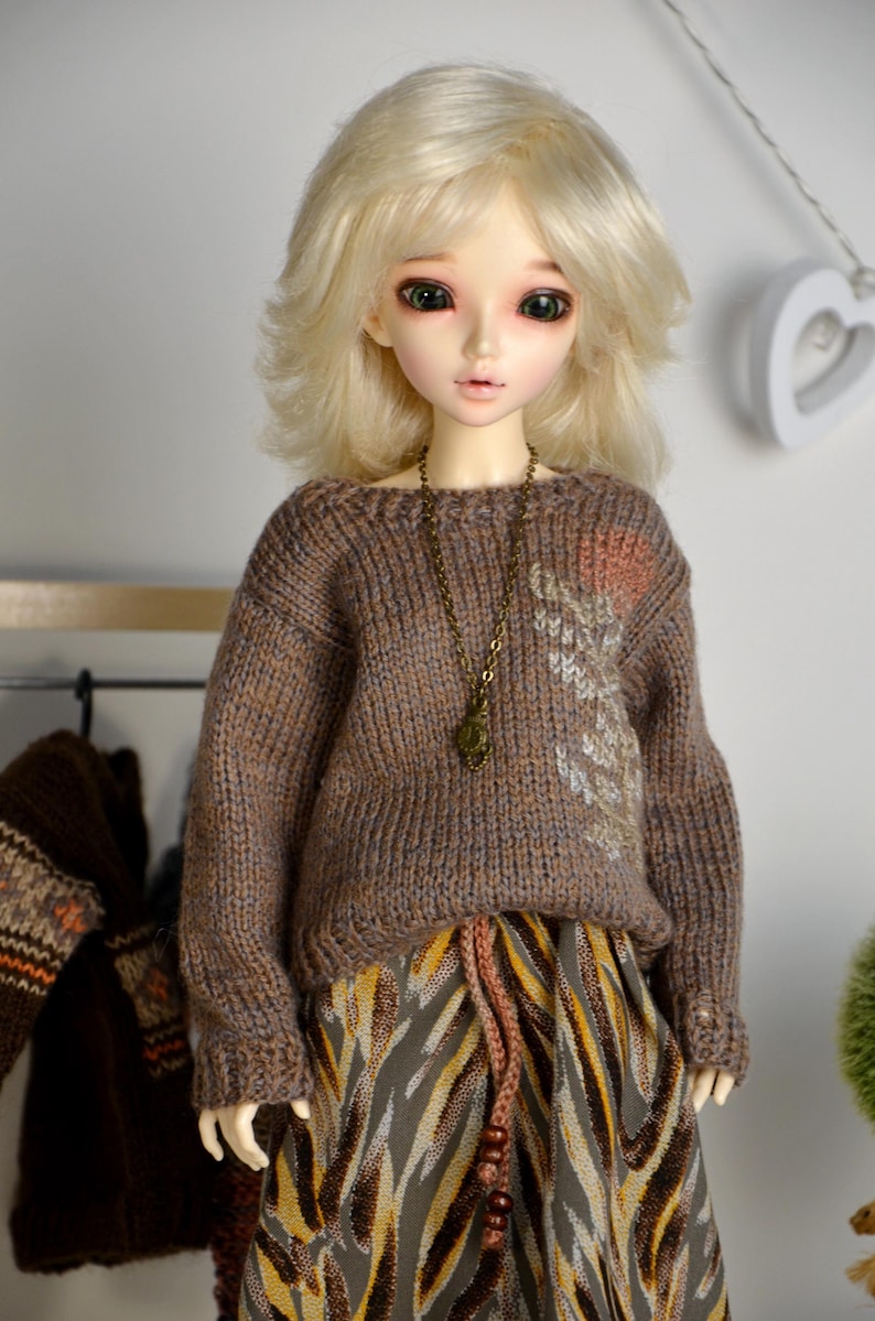 May include: A doll with blonde hair wearing a brown knit sweater with a decorative design and a patterned skirt. The doll has a long bronze necklace. The doll is standing in front of a white wall.
