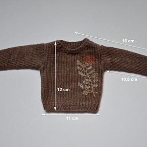 May include: A brown knitted sweater with a floral design. The sweater measures 12 cm in length, 11 cm in width, and the sleeves are 16 cm long. The sleeves are 10.5 cm long from the armpit to the cuff.
