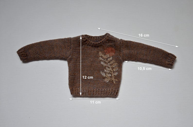 May include: A brown knitted sweater with a floral design. The sweater measures 12 cm in length, 11 cm in width, and the sleeves are 16 cm long. The sleeves are 10.5 cm long from the armpit to the cuff.