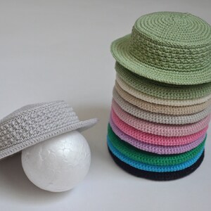 Peut inclure: Une pile de 10 chapeaux en crochet de diff&eacute;rentes couleurs, dont le vert, le beige, le rose, le violet, le bleu et le noir. Un seul chapeau en crochet gris se trouve sur le c&ocirc;t&eacute; gauche de l'image.