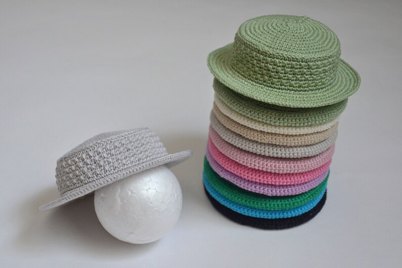 Peut inclure: Une pile de 10 chapeaux en crochet de diff&eacute;rentes couleurs, dont le vert, le beige, le rose, le violet, le bleu et le noir. Un seul chapeau en crochet gris se trouve sur le c&ocirc;t&eacute; gauche de l'image.
