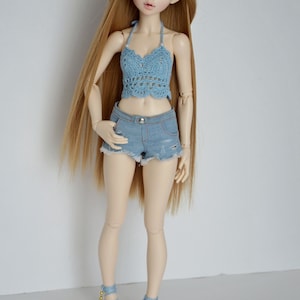 May include: A doll with long blonde hair wearing a light blue crochet halter top, denim shorts, and matching blue shoes. The doll's outfit has a summer theme, with a focus on handmade clothing and accessories.