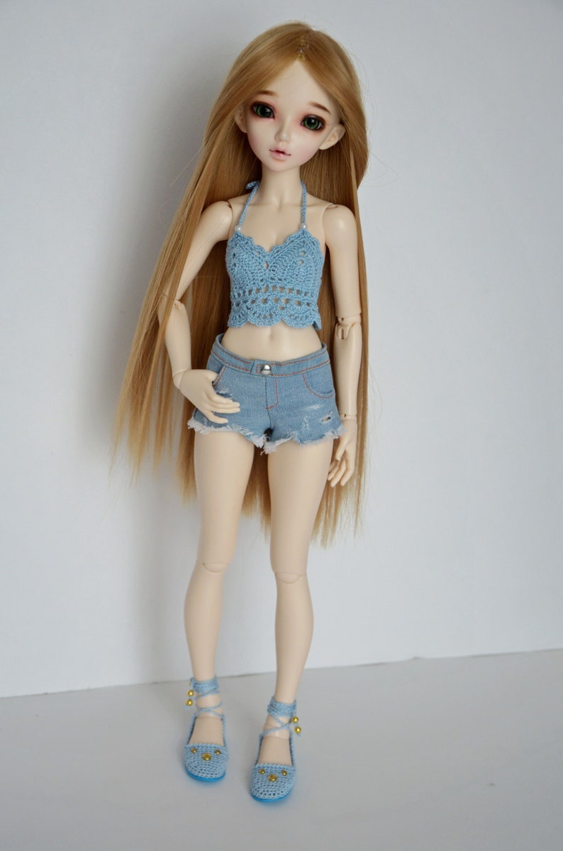 May include: A doll with long blonde hair wearing a light blue crochet halter top, denim shorts, and matching blue shoes. The doll's outfit has a summer theme, with a focus on handmade clothing and accessories.