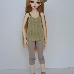 Peut inclure: Une poup&eacute;e aux longs cheveux blonds portant un chapeau en tricot vert, un d&eacute;bardeur en tricot vert, un pantalon marron et des chaussures en crochet marron.