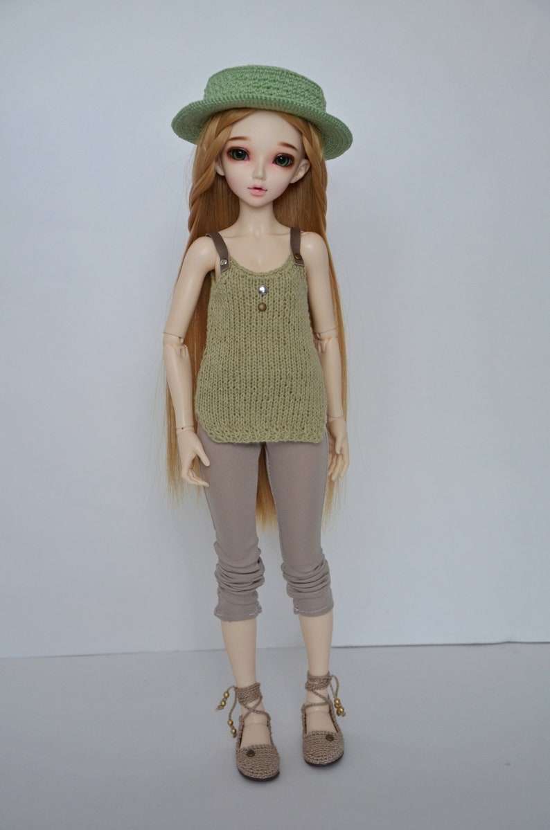 Peut inclure: Une poup&eacute;e aux longs cheveux blonds portant un chapeau en tricot vert, un d&eacute;bardeur en tricot vert, un pantalon marron et des chaussures en crochet marron.