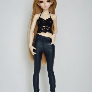 May include: A doll wearing a black crochet halter top and dark blue faux leather pants. The doll has long blonde hair and is standing against a white background.