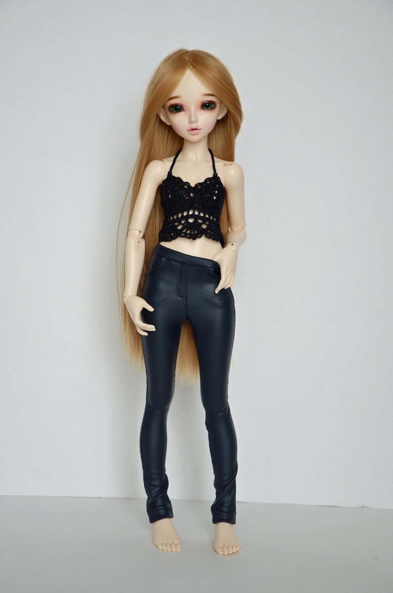 May include: A doll wearing a black crochet halter top and dark blue faux leather pants. The doll has long blonde hair and is standing against a white background.