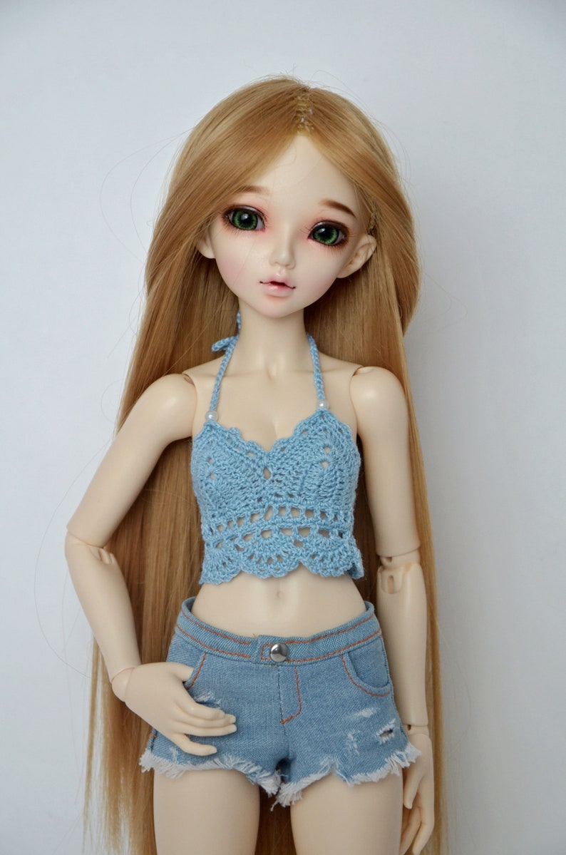 May include: A doll with long, light brown hair and green eyes wears a light blue crochet halter top and denim shorts. The shorts have a button closure and distressed detailing. The doll is posed against a white background.