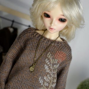 May include: A doll with pale skin and blonde hair wearing a brown knit sweater with a floral design and a bronze-colored necklace. The doll's sweater has a detailed pattern and a small clock pendant.