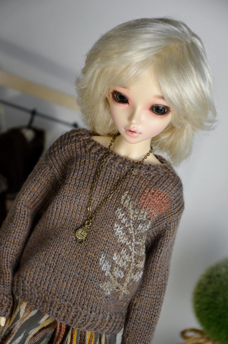 May include: A doll with pale skin and blonde hair wearing a brown knit sweater with a floral design and a bronze-colored necklace. The doll's sweater has a detailed pattern and a small clock pendant.
