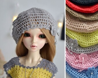Peut inclure: Un béret gris en crochet avec un embellissement floral et des perles argentées. Le béret est présenté sur une poupée aux cheveux blonds et aux yeux verts. L'image montre également une variété d'autres couleurs du même béret.