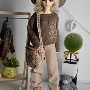 May include: A doll wearing a brown knitted hat, sweater, and matching purse. The doll is also wearing tan pants and boots. The sweater has a floral design. A small teddy bear and decorative items are in the background.
