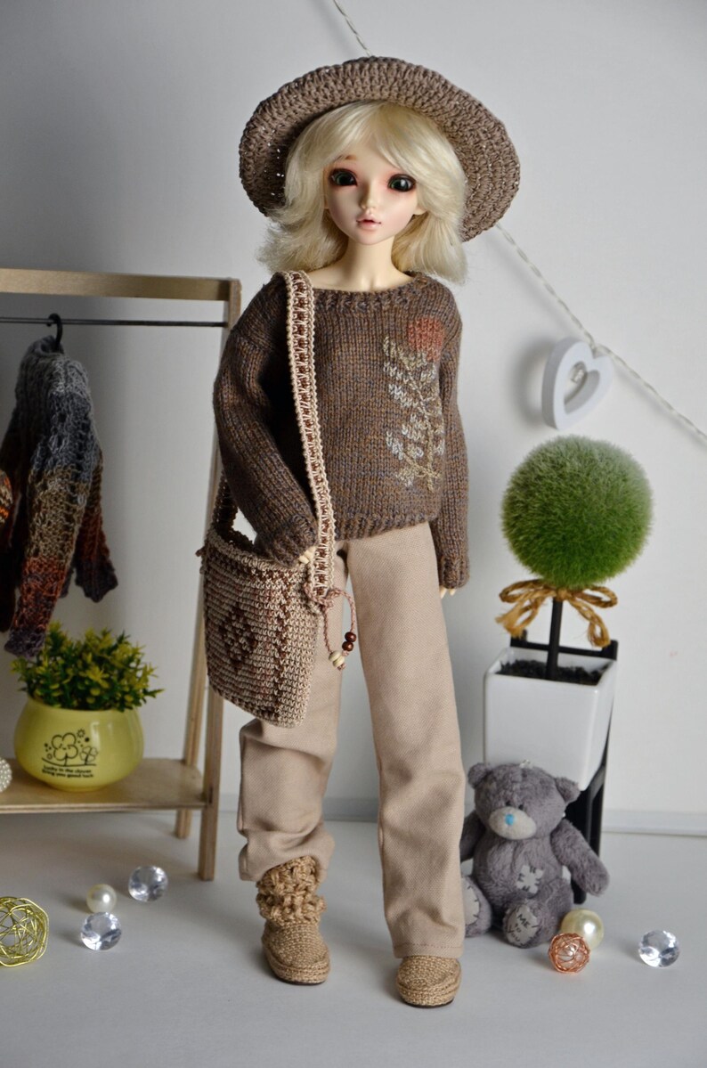 May include: A doll wearing a brown knitted hat, sweater, and matching purse. The doll is also wearing tan pants and boots. The sweater has a floral design. A small teddy bear and decorative items are in the background.