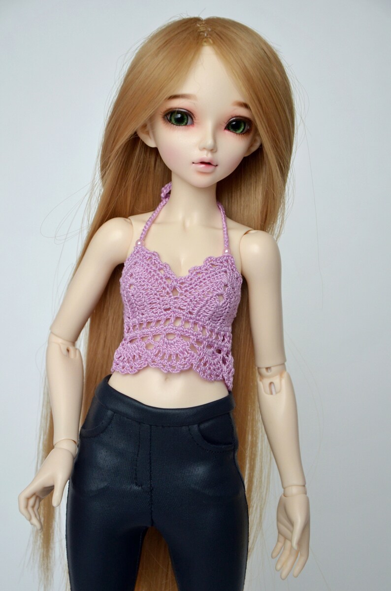 May include: A doll with long, light brown hair and green eyes is wearing a lavender crochet halter top and black pants. The doll is posed against a plain white background.