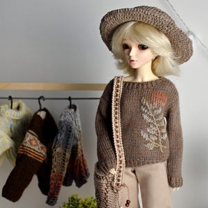 May include: A doll wearing a brown knit sweater with a floral design, a matching hat, and a shoulder bag. Several sweaters hang on a rack in the background. The doll is wearing tan pants.