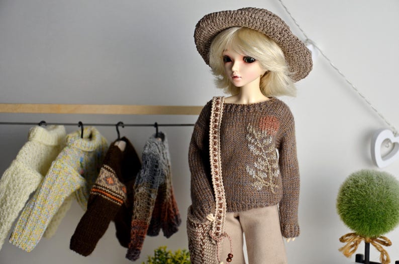 May include: A doll wearing a brown knit sweater with a floral design, a matching hat, and a shoulder bag. Several sweaters hang on a rack in the background. The doll is wearing tan pants.