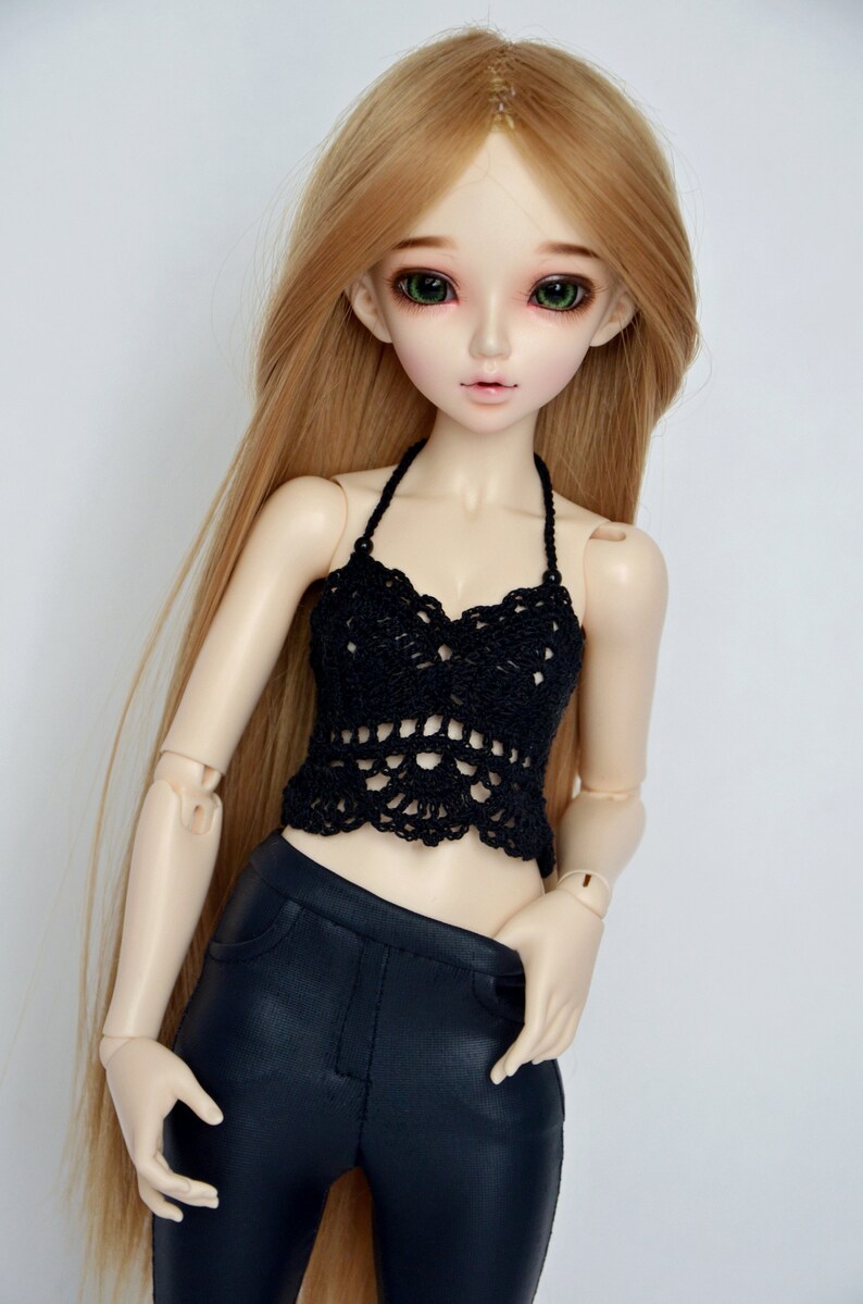 May include: A doll with long blonde hair and green eyes wearing a black crochet halter top and black pants. The doll has a pale complexion and is posed against a white background. The doll's clothing is detailed.