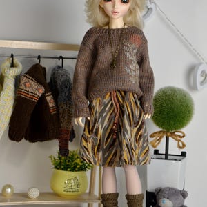 May include: A doll wearing a brown sweater with a leaf design, a patterned skirt, and brown crochet boots. A rack of miniature sweaters hangs in the background. A small teddy bear and decorative items are also present.
