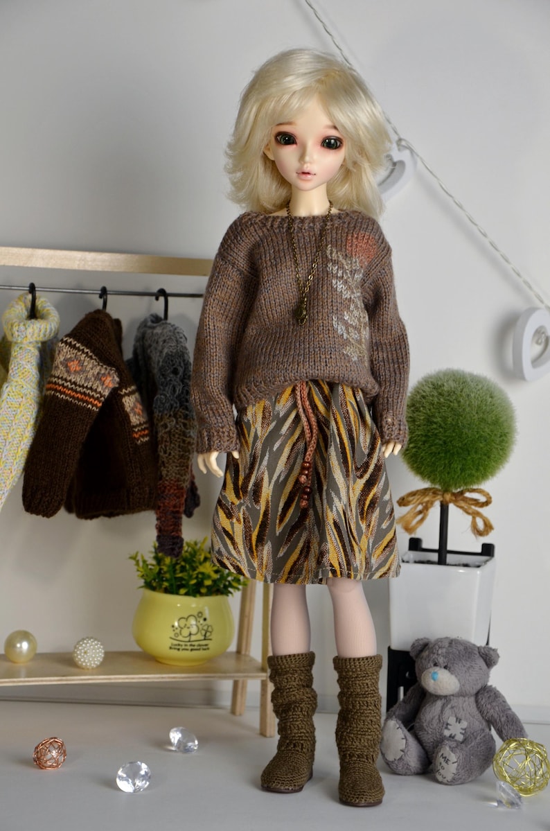 May include: A doll wearing a brown sweater with a leaf design, a patterned skirt, and brown crochet boots. A rack of miniature sweaters hangs in the background. A small teddy bear and decorative items are also present.