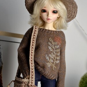 May include: A doll wearing a brown knitted hat, sweater, and a matching shoulder bag. The sweater features an embroidered floral design. The doll is also wearing blue jeans. The doll has blonde hair and is posed in front of a neutral background.
