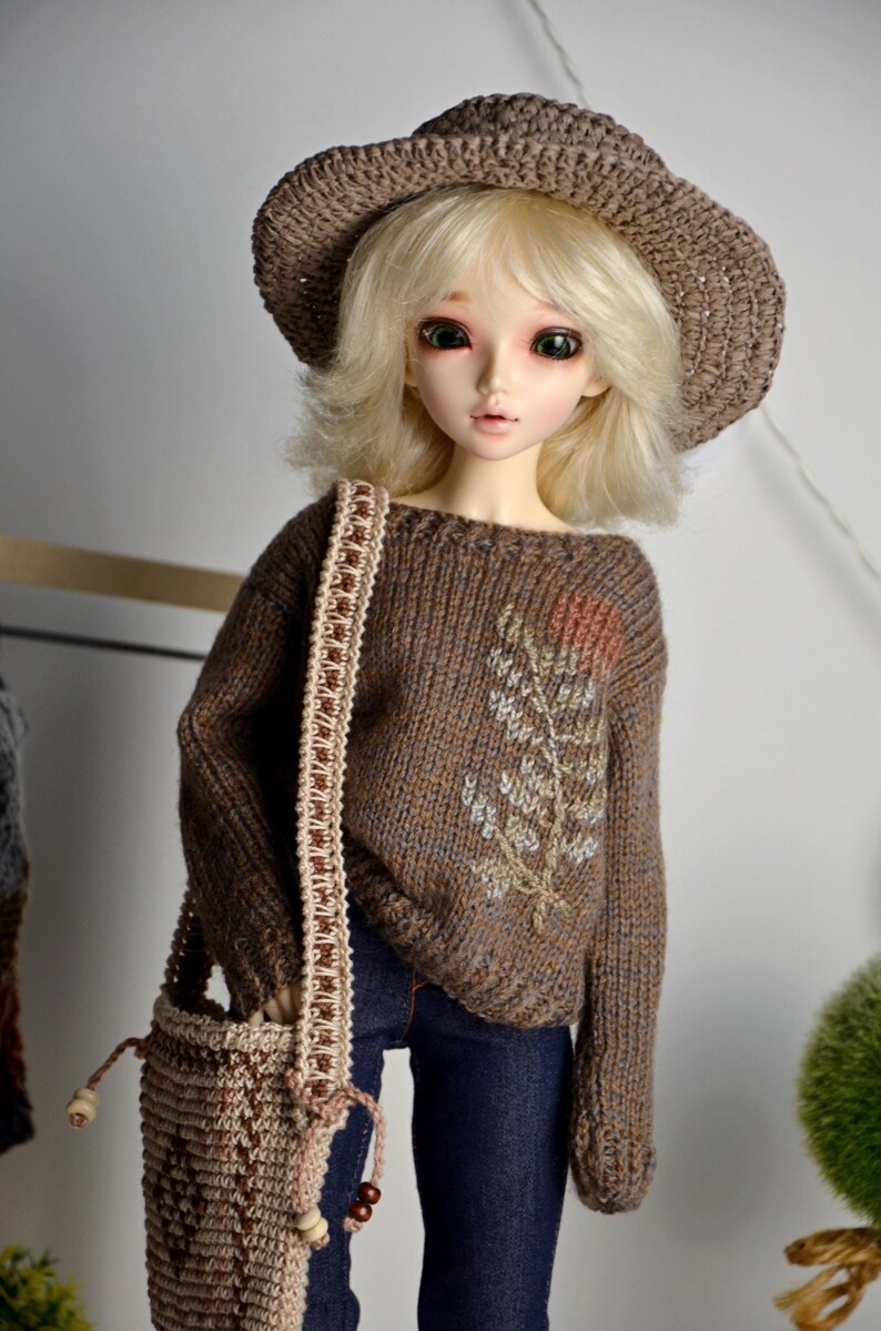 May include: A doll wearing a brown knitted hat, sweater, and a matching shoulder bag. The sweater features an embroidered floral design. The doll is also wearing blue jeans. The doll has blonde hair and is posed in front of a neutral background.
