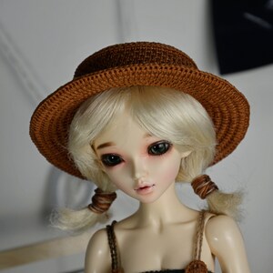 Peut inclure: Une poup&eacute;e aux cheveux blonds portant un chapeau en crochet marron. La poup&eacute;e a les yeux verts et porte un bandeau en crochet marron.