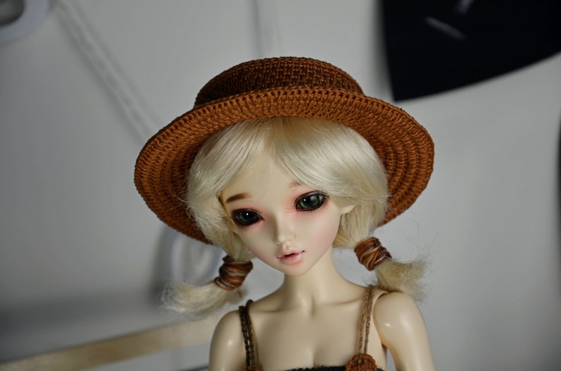Peut inclure: Une poup&eacute;e aux cheveux blonds portant un chapeau en crochet marron. La poup&eacute;e a les yeux verts et porte un bandeau en crochet marron.