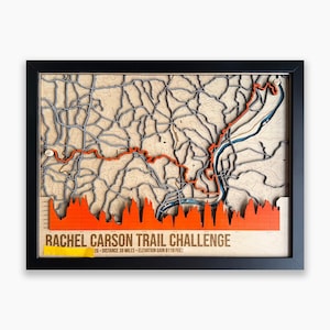 May include: A framed 3D wooden topographic map of the Rachel Carson Trail Challenge. The map features a light wood background with grey terrain lines, a bright orange trail path, and an orange elevation profile graph with 38 miles and 8119 feet.