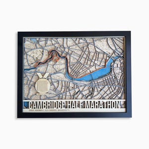 Op de afbeelding: Houten kaart van Cambridge, Massachusetts, in een lijst, met een blauwe rivier en een rood omlijnd pad. Een medaille staat linksonder, met de tekst "CAMBRIDGE HALF MARATHON" onderaan.